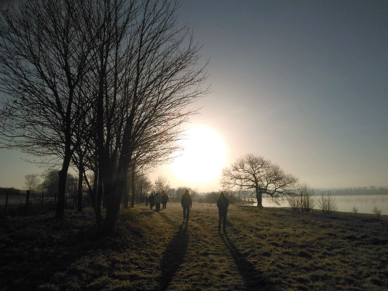 Deux marcheurs à contre-jour, au lever du soleil, près d’un plan d’eau.