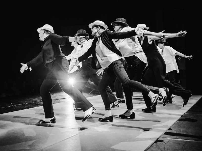Des danseurs de claquettes en chapeau s’élancent en file, bras tendus. Photo en noir et blanc.